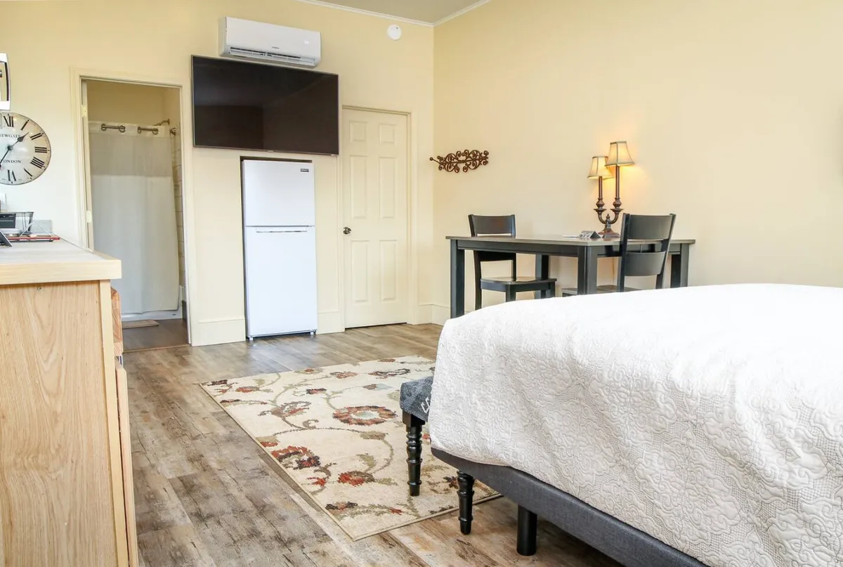 A bright, comfortable living area with a Queen-size bed, a small dining table set for two, a refrigerator, a wall-mounted TV, and a lamp. The space is welcoming, designed for both relaxation and connection, with the TV positioned conveniently for entertainment from bed or table.