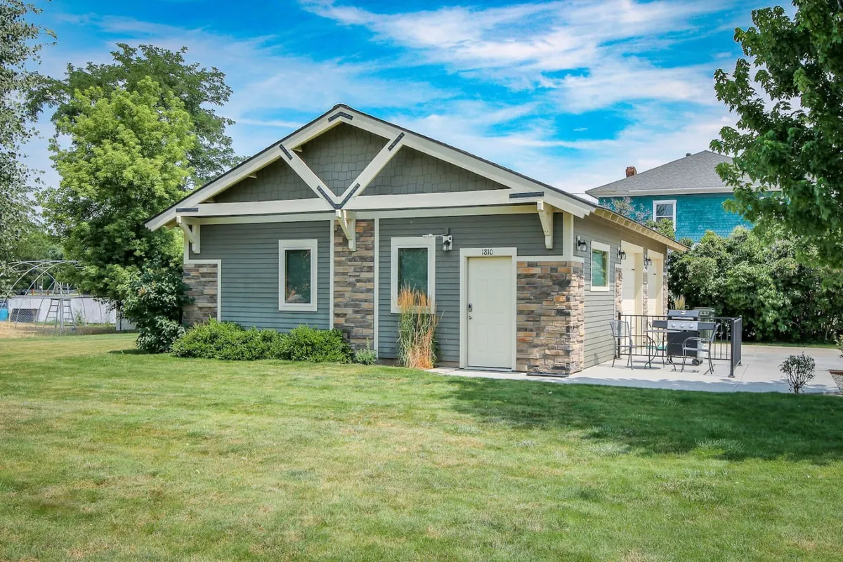 The image showcases the exterior of a charming studio cottage with a classic stone facade, an inviting front door, a tastefully landscaped yard, and a cozy patio area perfect for outdoor dining and relaxing. The mood is welcoming, with lush greenery and comfortable seating that hint at both relaxation and opportunities for outdoor gatherings.