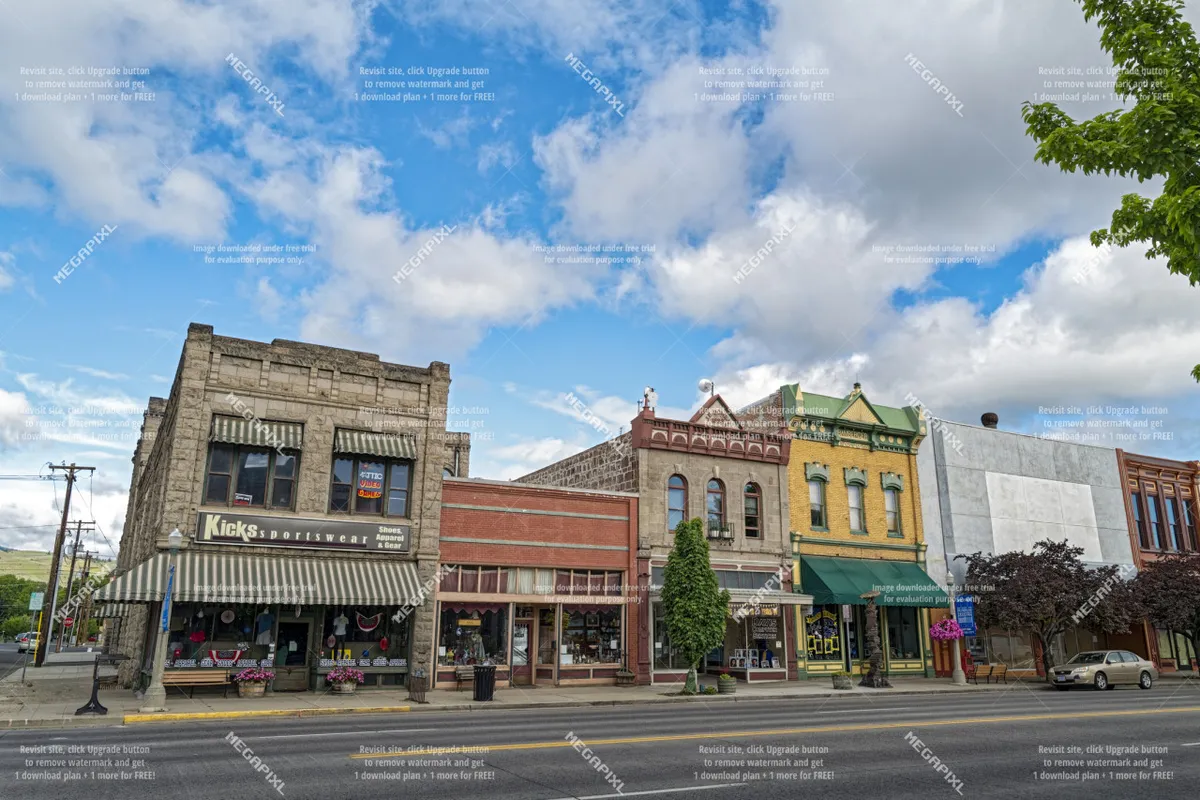 A bustling streetscape scene with historic buildings, boutique storefronts, and leafy street trees. The sidewalk is pedestrian-friendly, lined with shops and dining spots, capturing the vibrant energy of downtown Baker City.