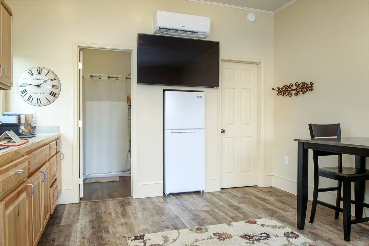 A cozy dining and living area with wooden cabinetry, a large wall clock, a flat-screen TV, refrigerator, and a small dining table. The space is set for shared meals, movie nights, and comfortable downtime after a day of exploring.