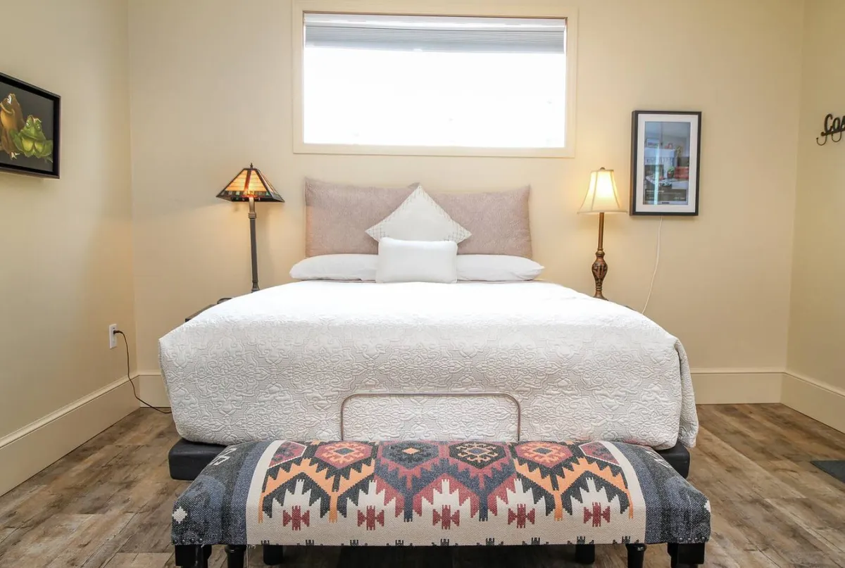 A cozy interior space featuring a plush Queen-size bed adorned with soft bedding, a decorative bench at the foot, warmly lit table lamps, and tasteful wall artwork. The atmosphere is serene and inviting, ideal for relaxation or unwinding after a day out.