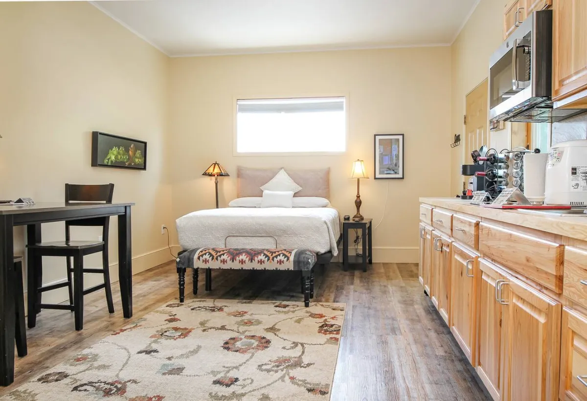An inviting interior that blends a bed with decorative pillows, a compact kitchenette, and a dining table with chairs, all on a patterned area rug. The scene evokes comfort and convenience—ideal for both rest and socializing within the private space.